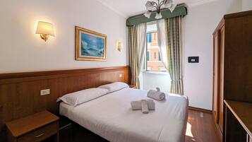 A hotel room with a wooden bed, a nightstand, a painting, a window with curtains, and a chandelier.