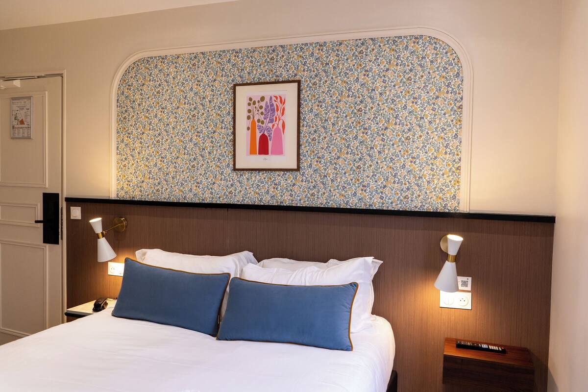 A hotel room with a neatly made bed, two bedside lamps, a framed artwork on the wall, and a wooden nightstand with a remote control.