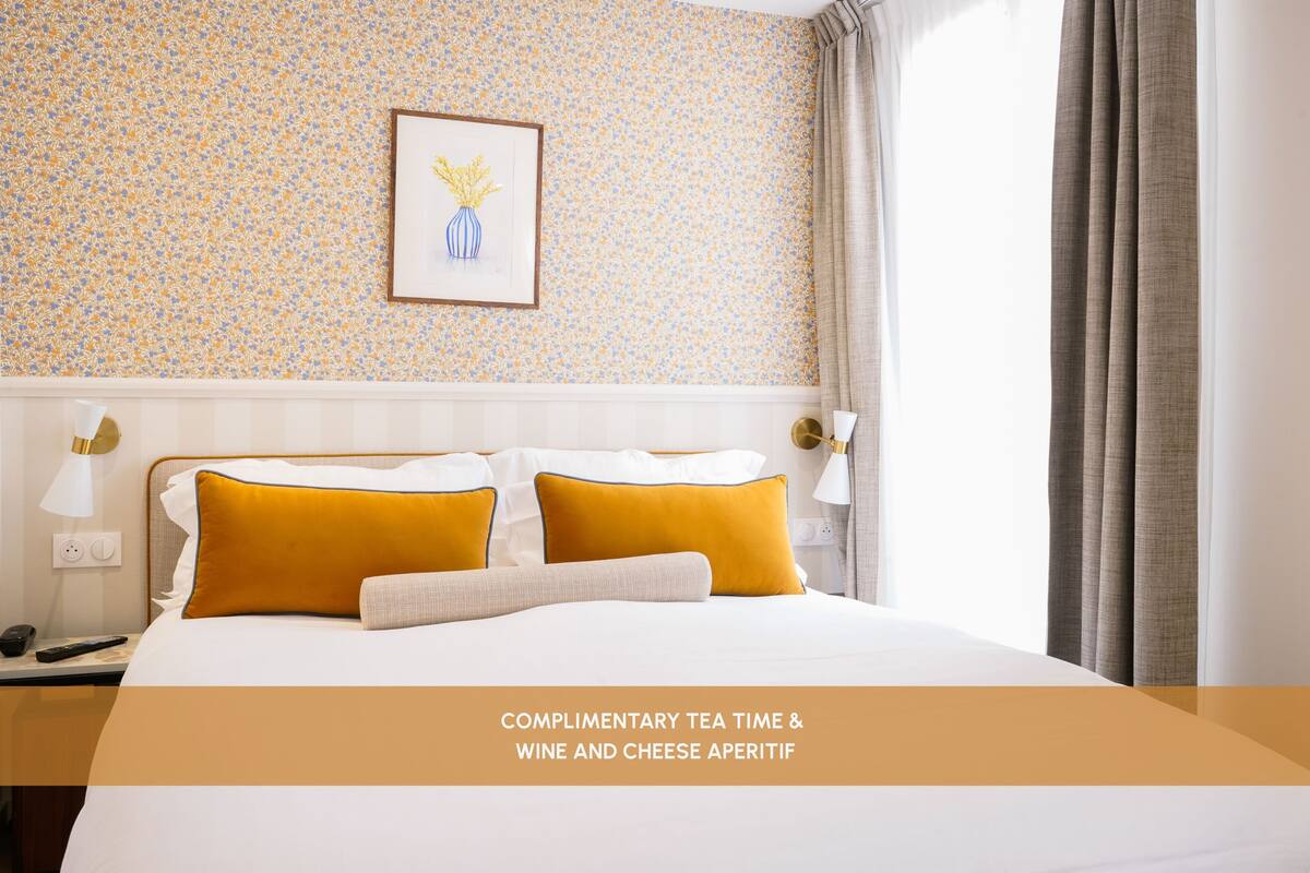 A hotel room with a bed, pillows, a nightstand, a lamp, a framed picture on the wall, and a window with curtains.