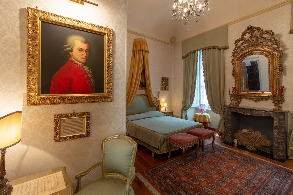 A bedroom with a bed, a chair, a mirror, a fireplace, and a painting on the wall.