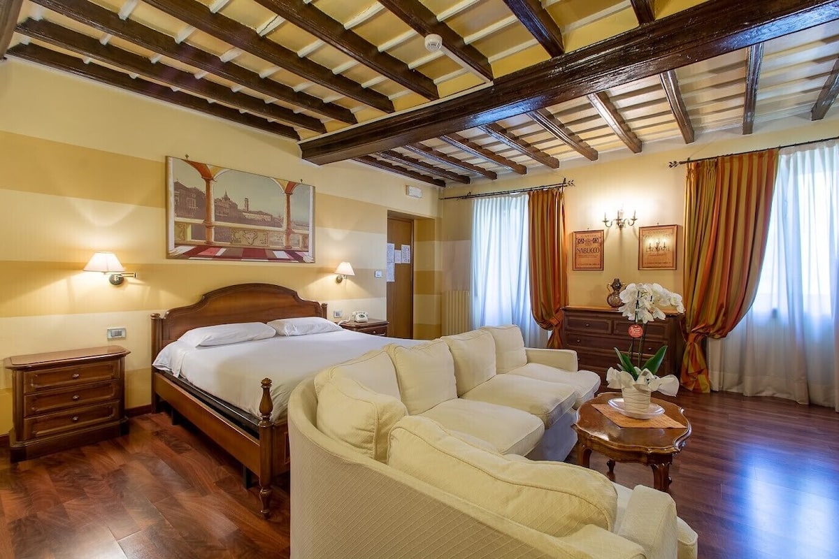 A hotel room with a wooden bed, a sofa, a nightstand, a lamp, and a painting on the wall.