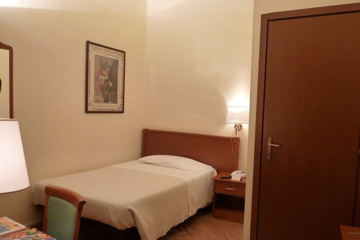 A small, neatly arranged bedroom with a single bed, a bedside table with a lamp, a chair, and a door.