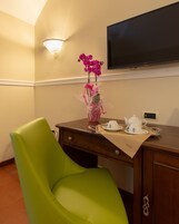 Una camera d'albergo con una poltrona verde, una scrivania con un vaso di orchidee rosa, una televisione e un angolo caffè.