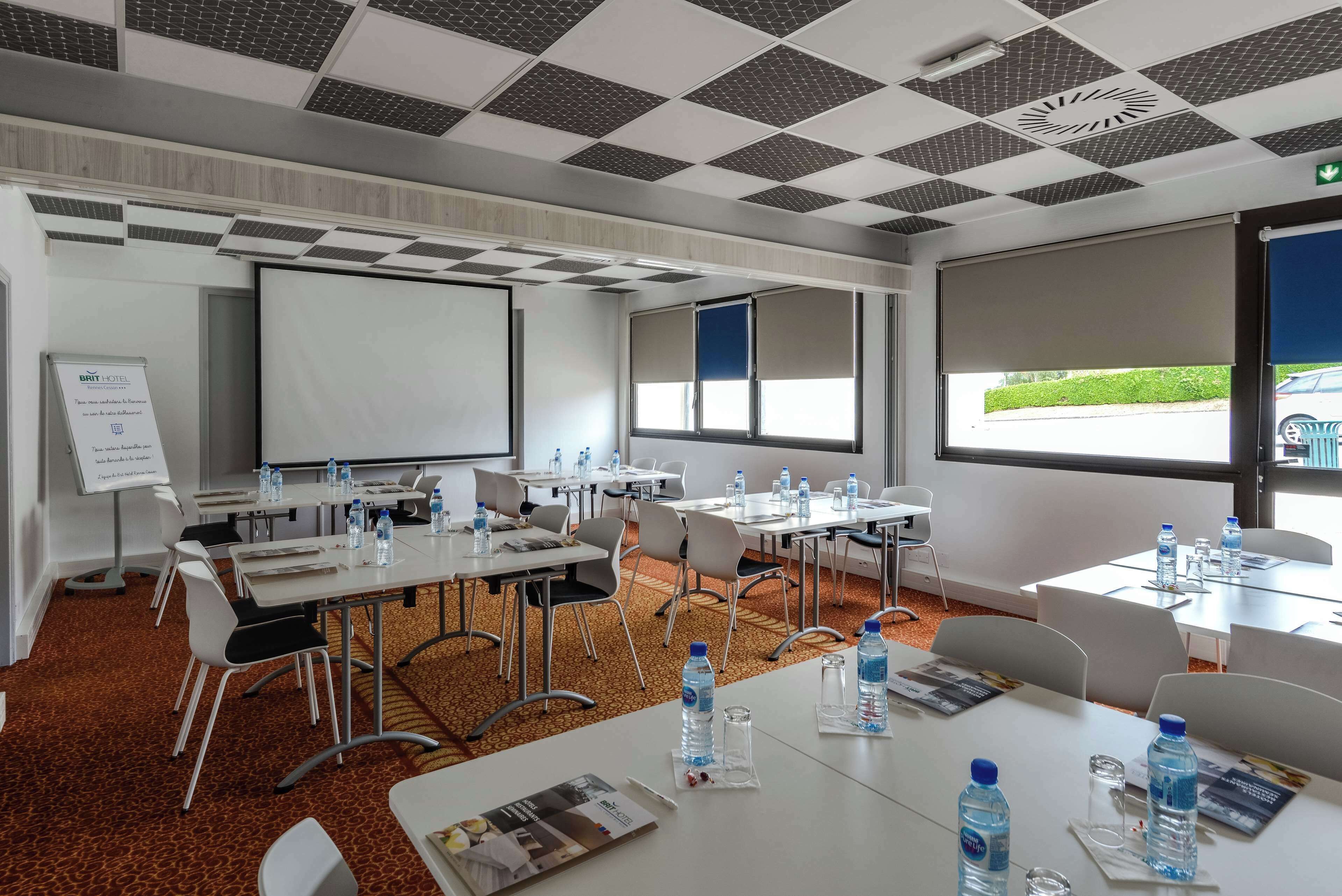 A conference room with white tables, chairs, and a projector screen. Water bottles and pamphlets are on the tables.
