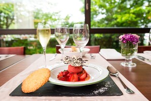 A dessert plate with strawberries and a chocolate garnish, accompanied by a bread-like item, on a table with wine glasses and a vase of flowers.