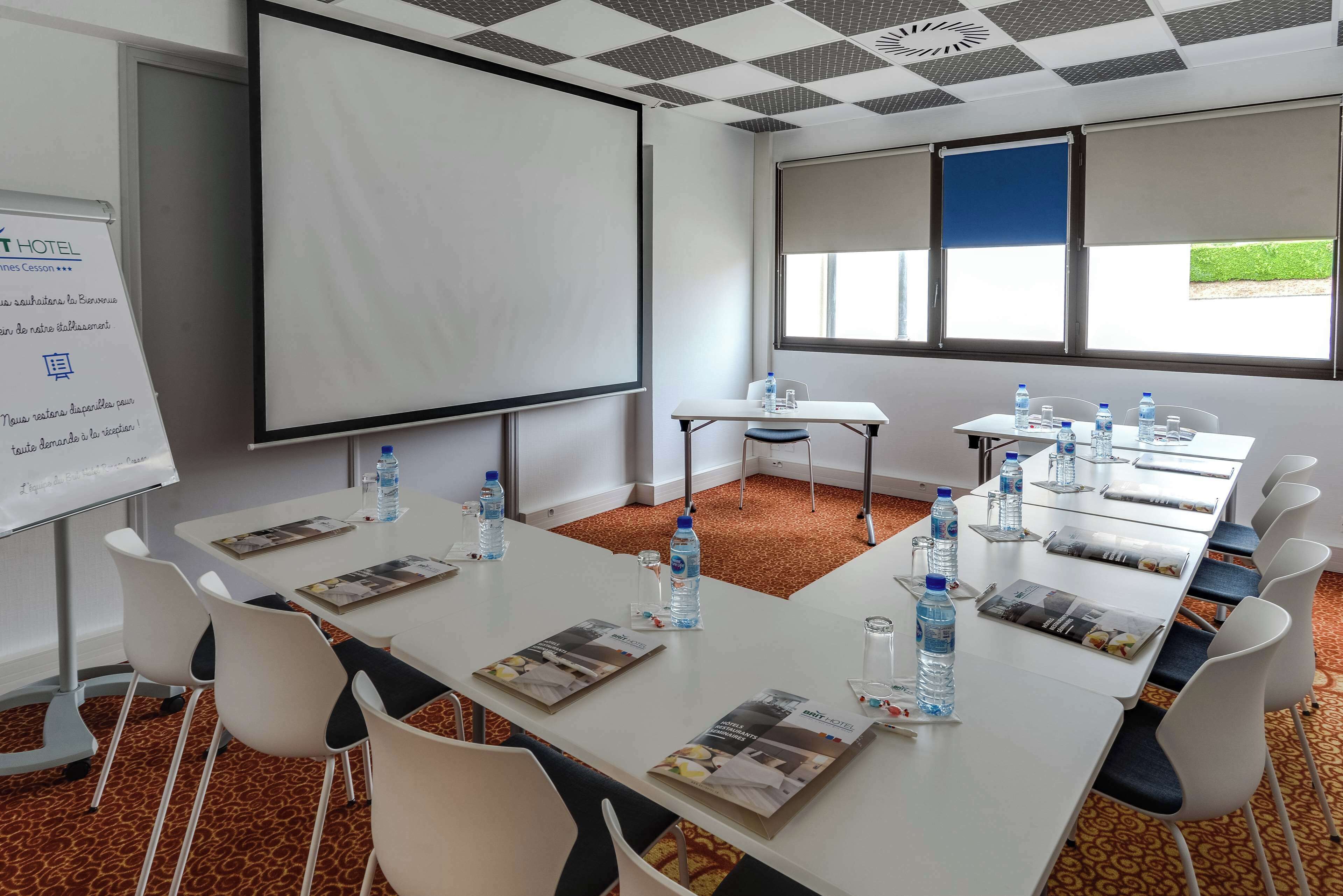 Un salón de conferencias con una pizarra, pantalla de proyector y varias mesas con botellas de agua y revistas.