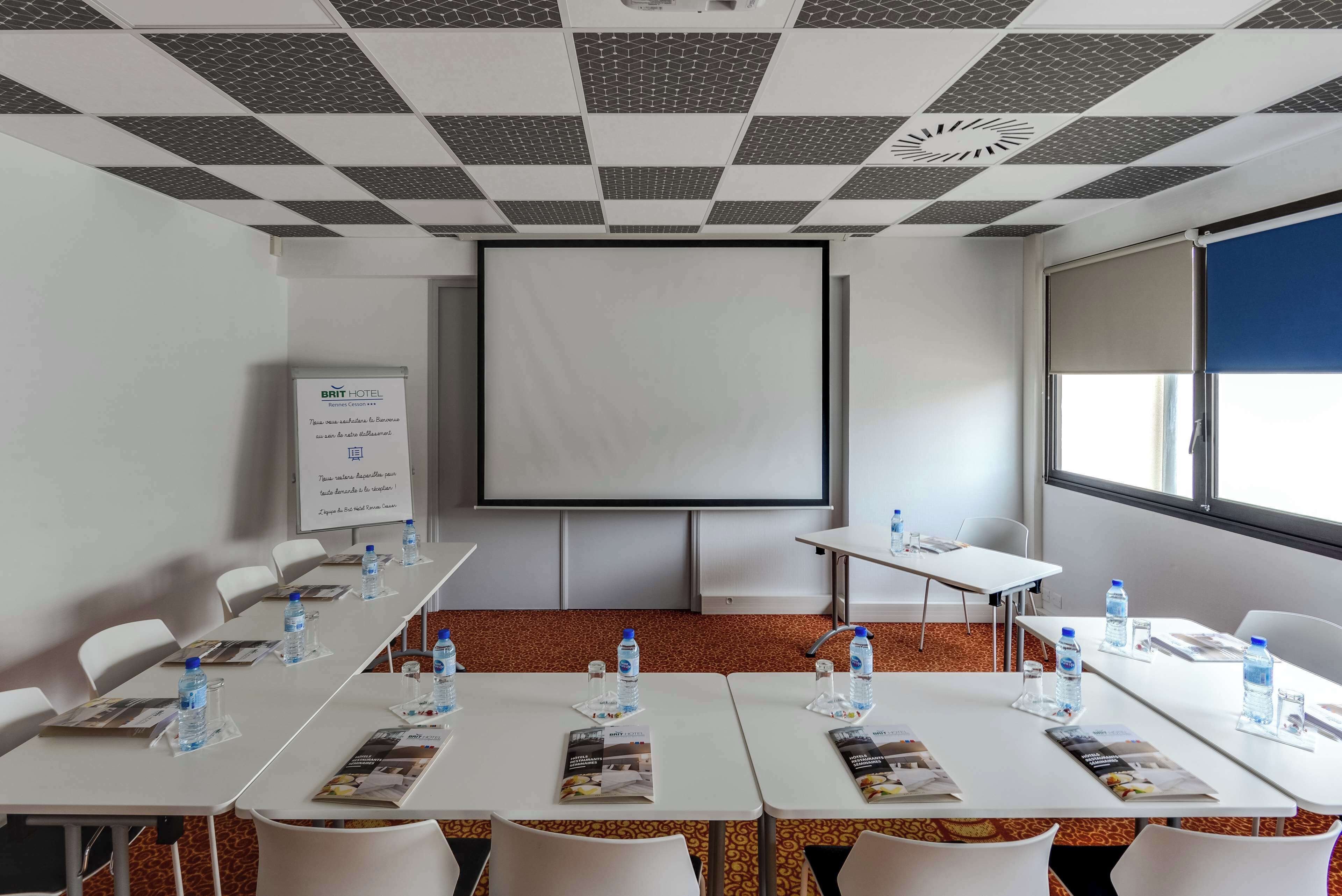 Un salón de conferencias con una pantalla grande, mesas blancas, sillas, botellas de agua y folletos informativos.