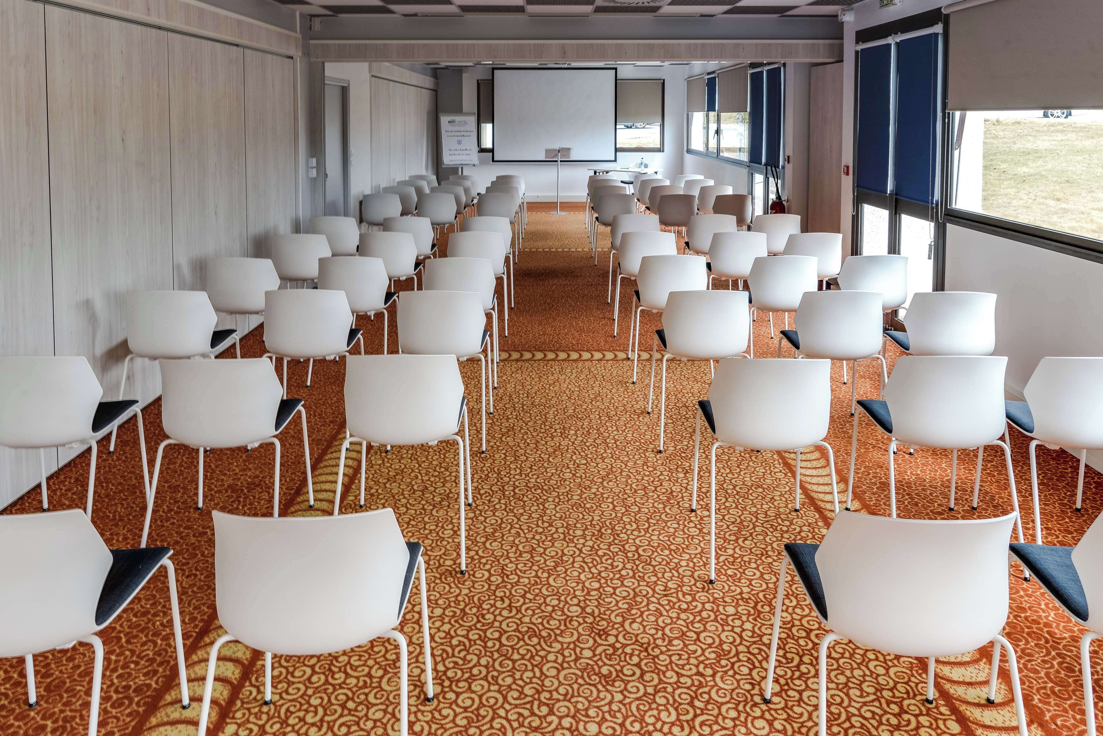 Un salón de conferencias con filas de sillas blancas, piso alfombrado, una pantalla grande y ventanas.