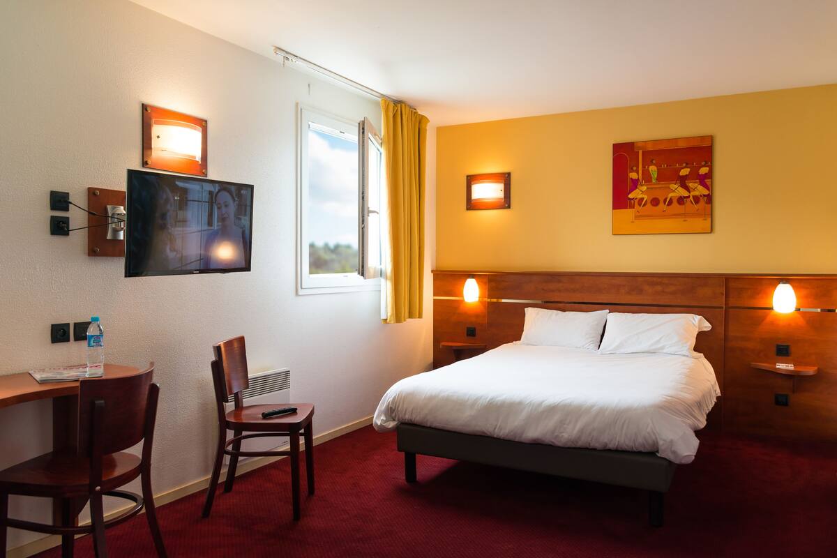 A hotel room with a bed, a desk with a chair, a television, and two wall-mounted lights.