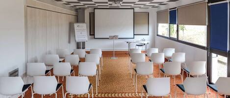 A conference room with white chairs arranged in rows, a carpeted floor, a projector screen, and a whiteboard.