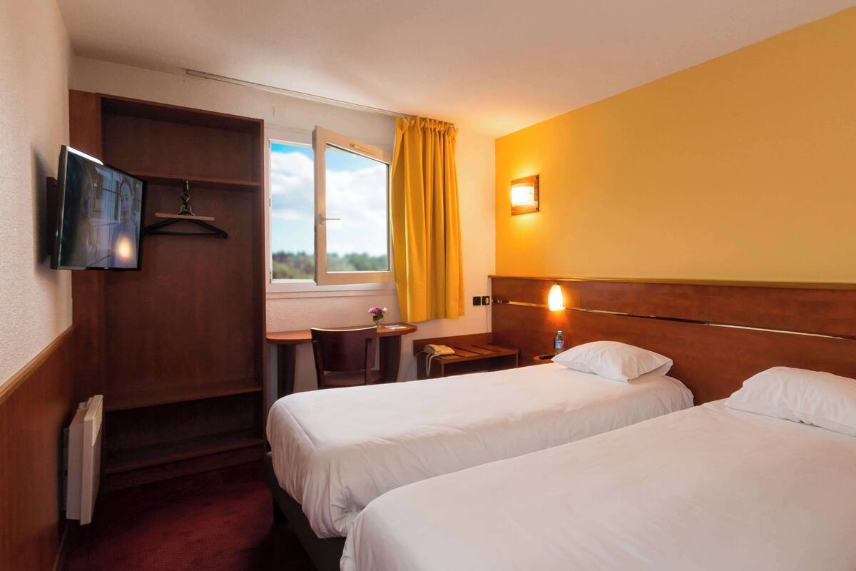 A hotel room with two beds, a desk, a chair, a small table, a TV mounted on the wall, and a window with curtains.