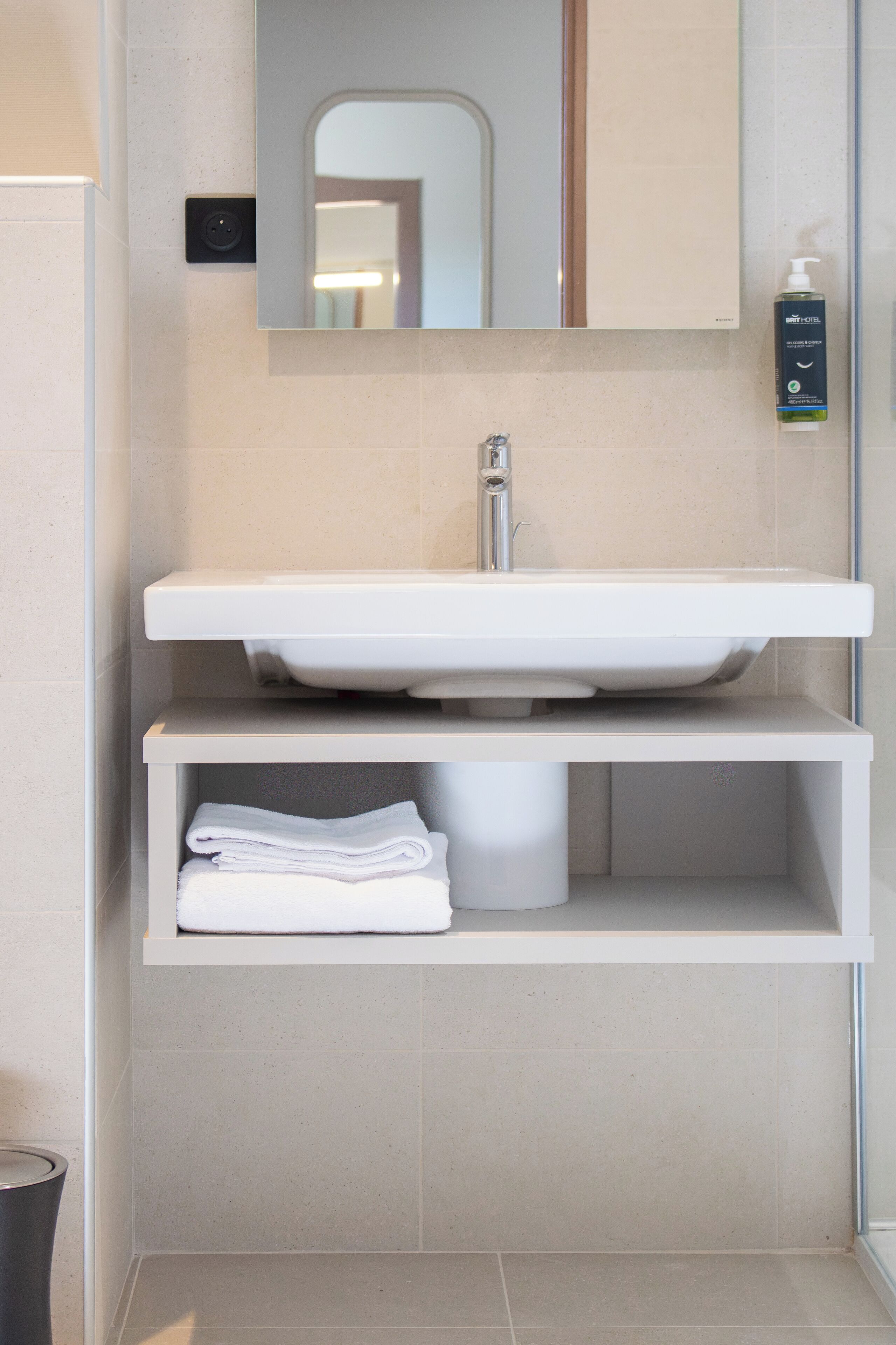 Ein Badezimmer mit einem weißen Waschbecken, einem Spiegel darüber, einem Regal mit gefalteten Handtüchern und einer Handseifenflasche.