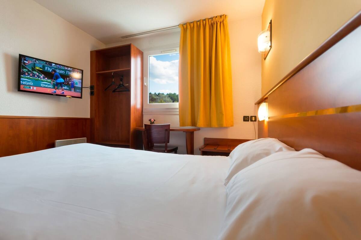 A hotel room with a bed, a desk, a TV, a window with curtains, and a wardrobe.
