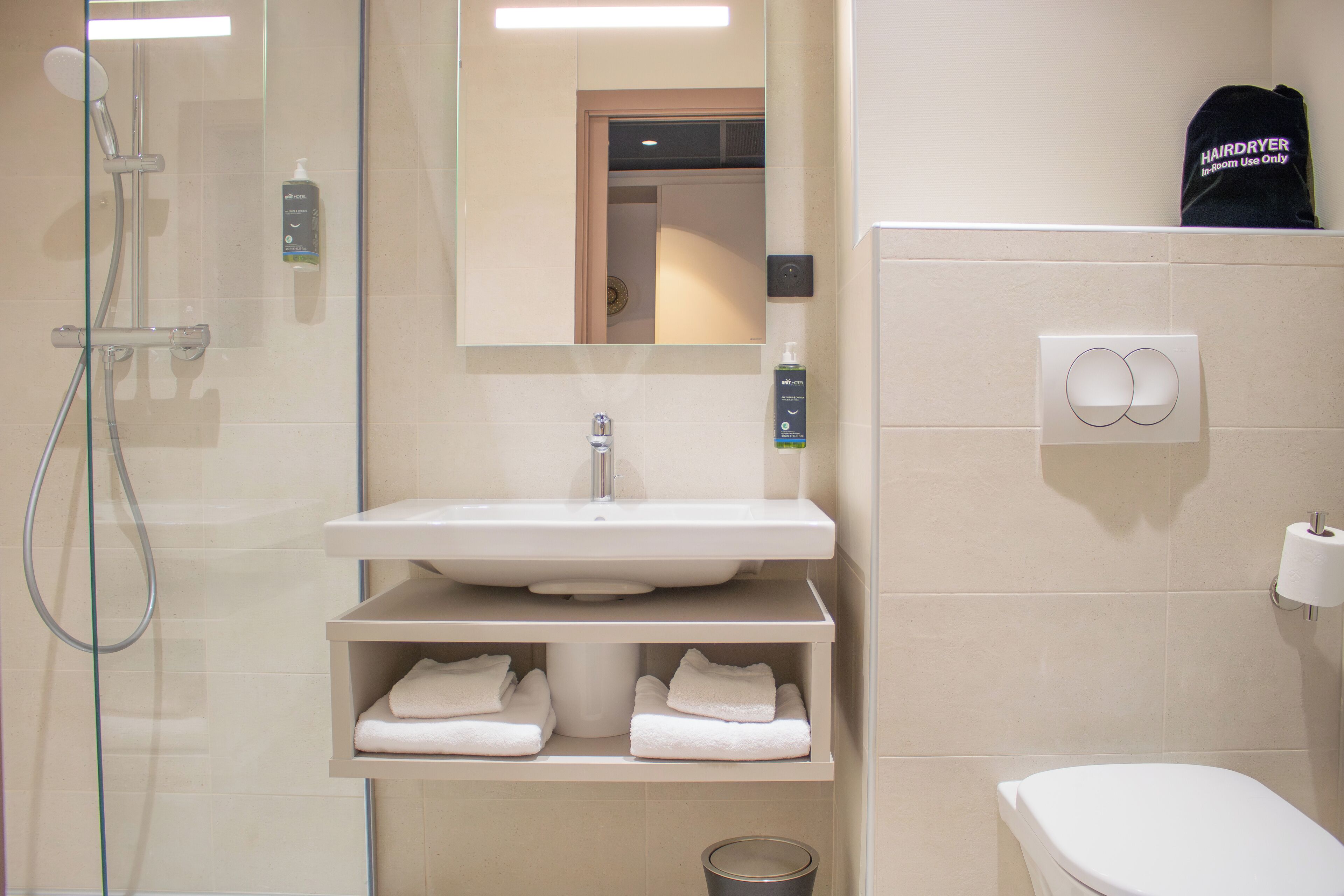 A bathroom with a glass shower enclosure, a sink with a mirror above it, and a toilet. There are towels on a shelf and a hairdryer on the wall.
