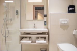 Et badeværelse med brusekabine i glas, en vask med et spejl over og et toilet. Der er håndklæder på en hylde og en hårtørrer på væggen.