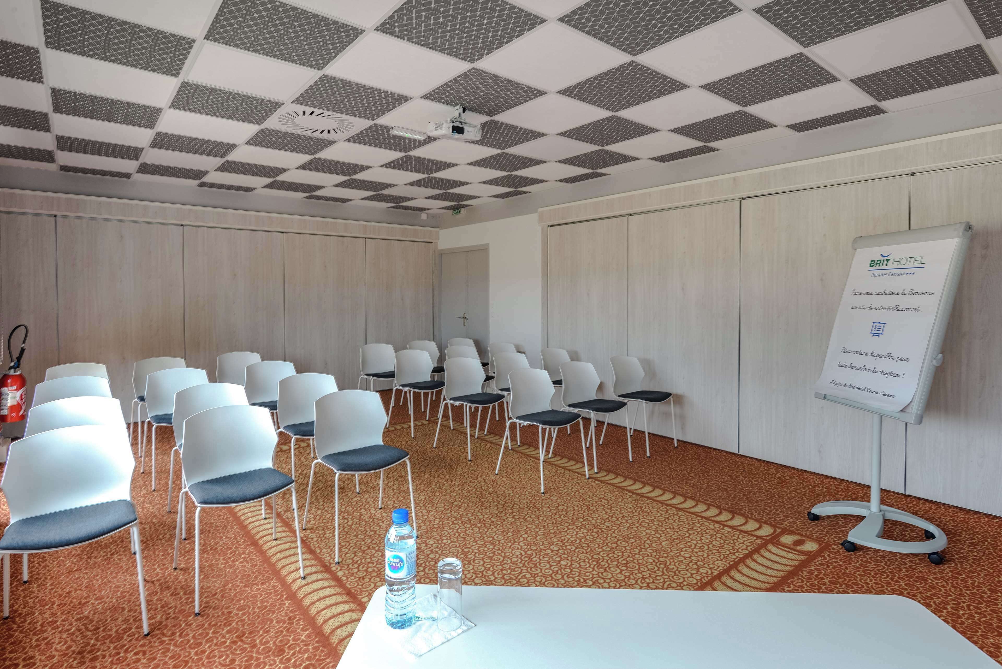 Sala de conferências com fileiras de cadeiras brancas, um quadro branco e piso carpeteado.