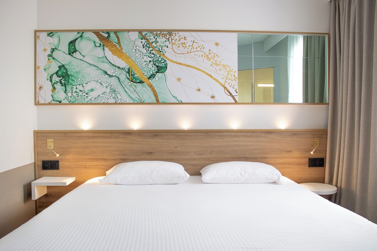 A hotel room with a large bed, a wooden headboard, bedside tables, a wall-mounted mirror, and a large abstract painting.