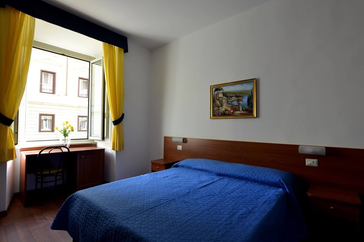 Habitación de hotel con cama, escritorio, silla, mesita de noche y un cuadro en la pared.