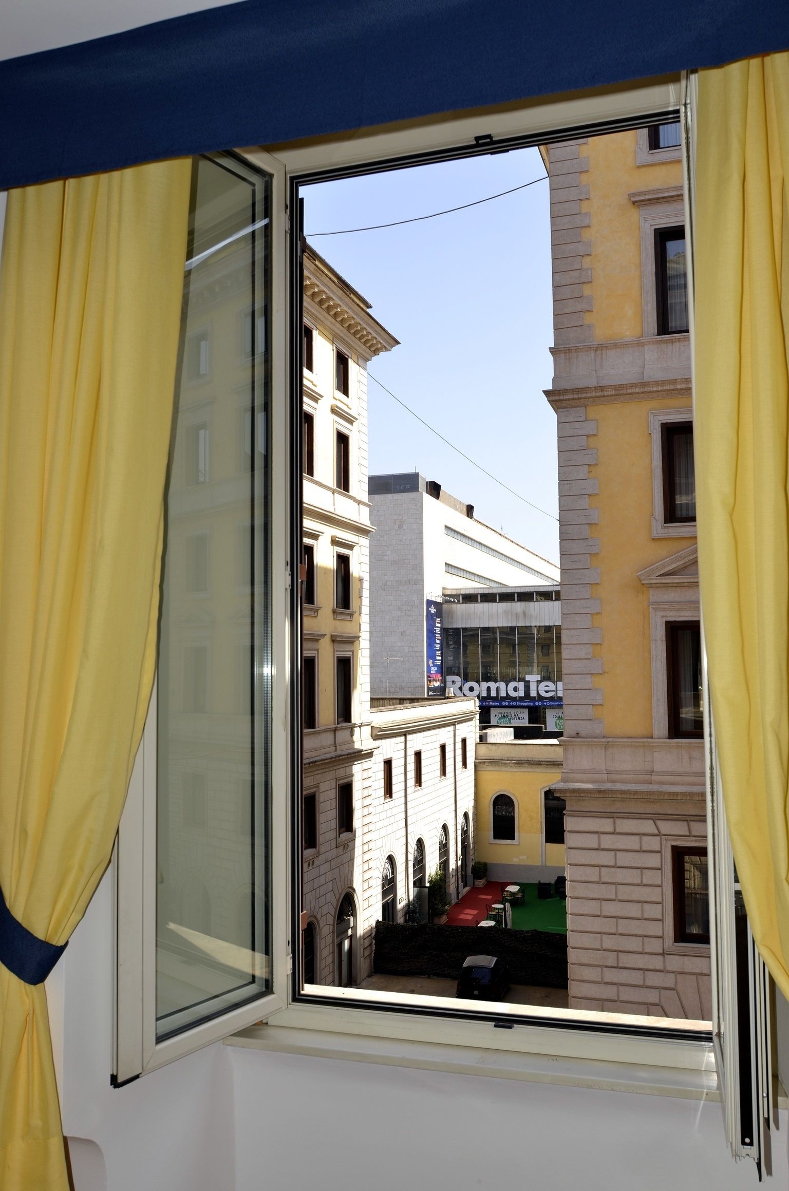 A window with yellow curtains, a view of a building with a 'RomaTei' sign, and a red carpeted area outside.