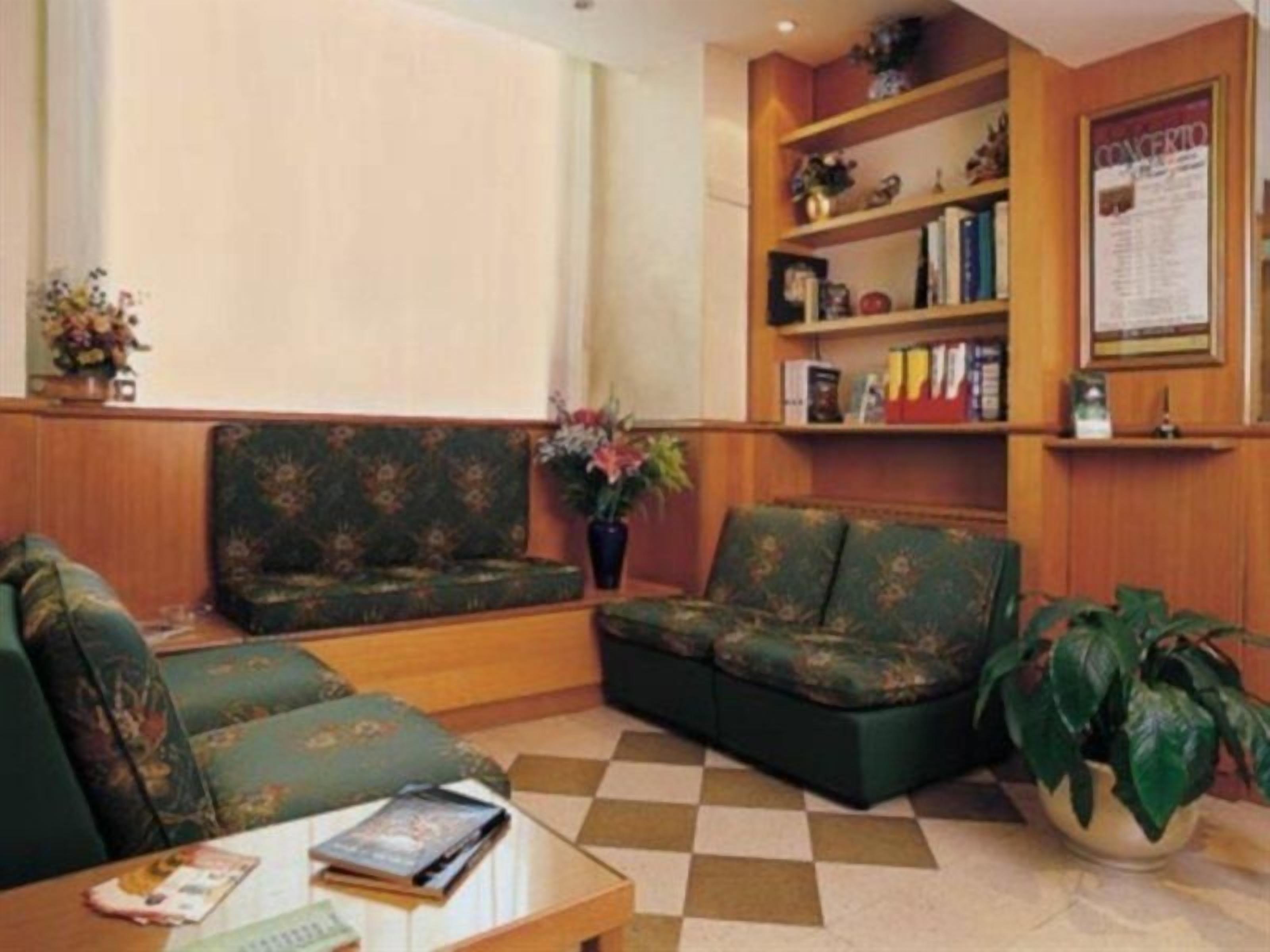 A lobby with a checkered floor, two green upholstered sofas, a coffee table with books, a bookshelf, and a potted plant.