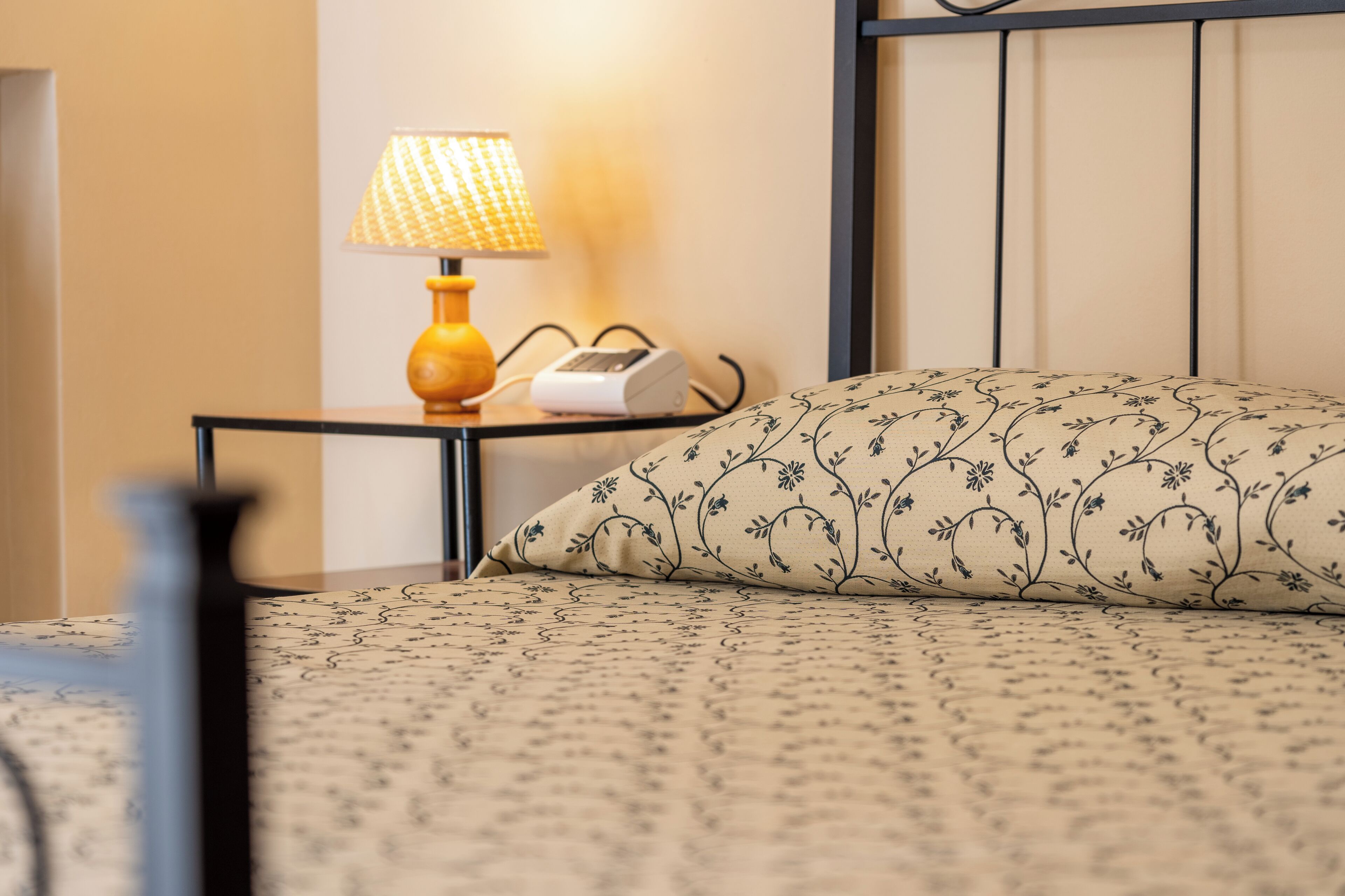 A neatly made bed with a floral-patterned pillow and a bedside table with a lamp and a device.