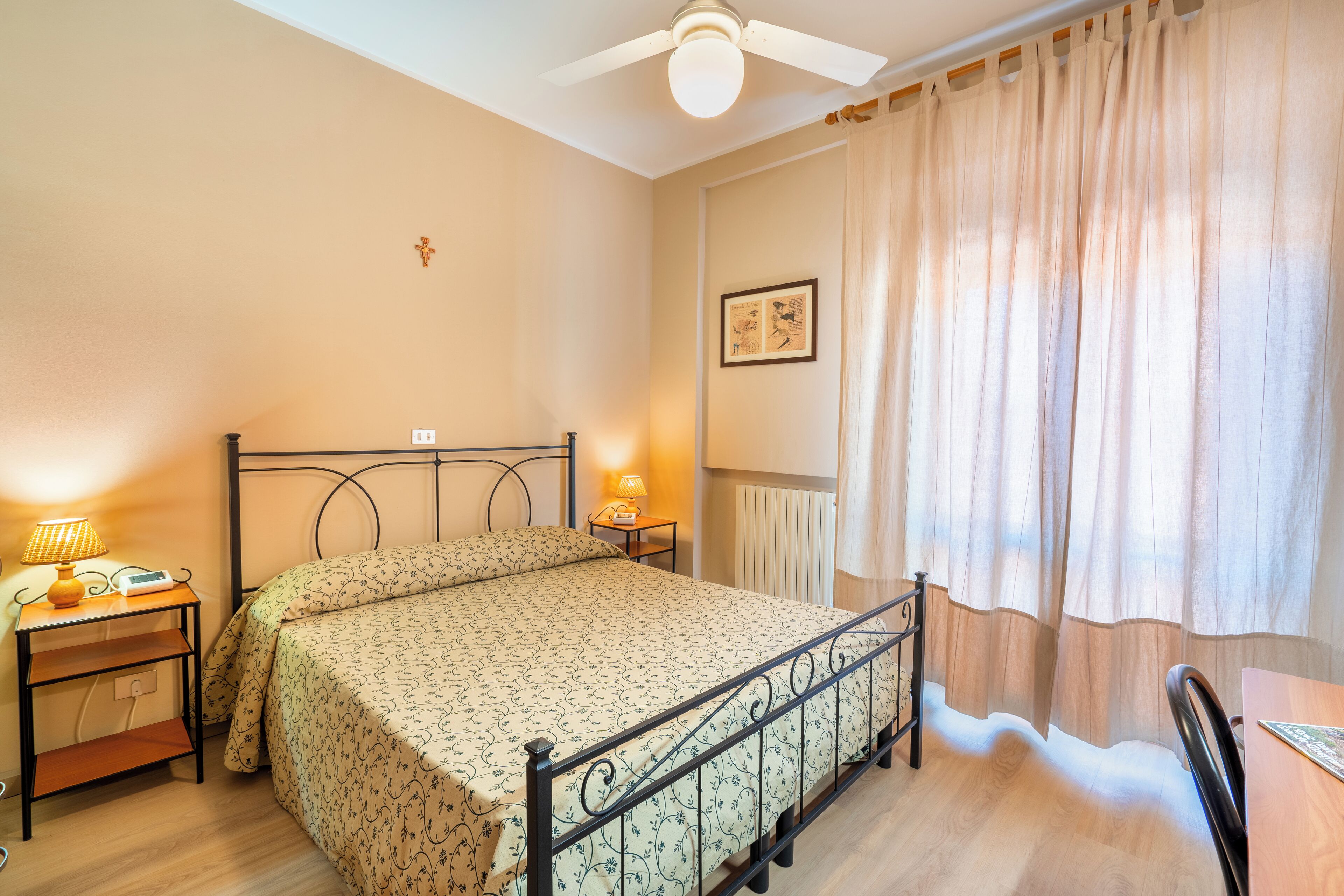 A bedroom with a bed, bedside tables, a ceiling fan, a radiator, and a window with curtains.