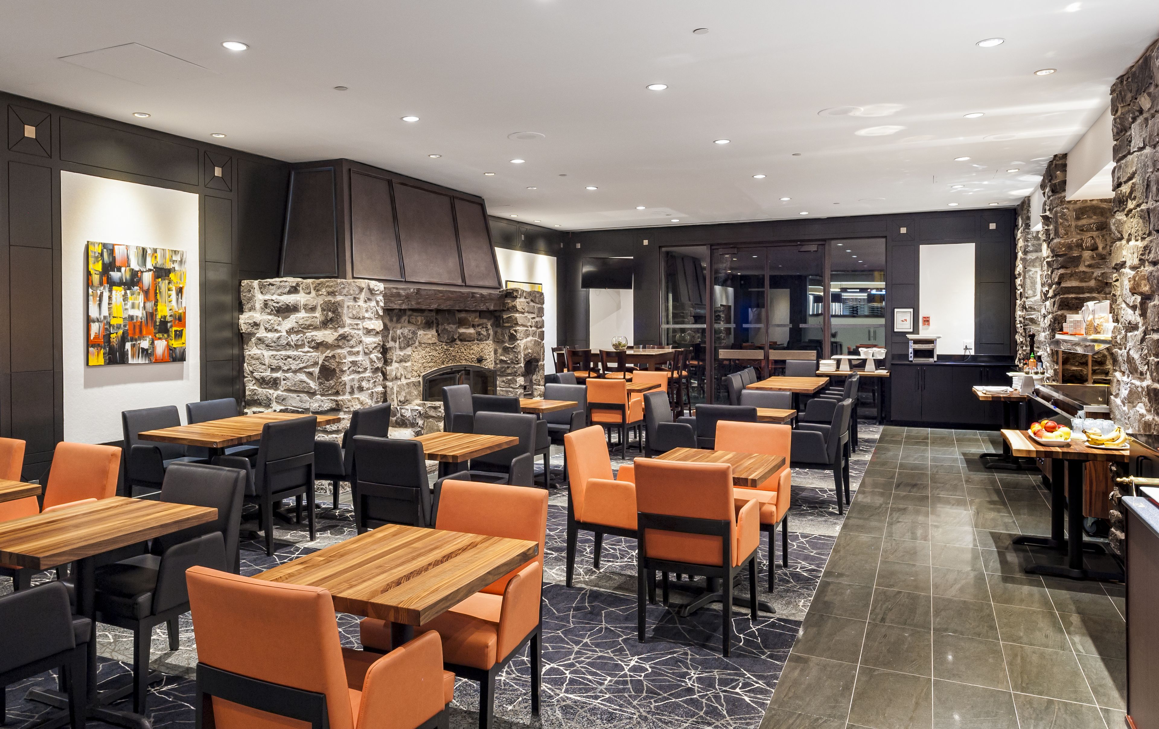 A modern restaurant with wooden tables and orange chairs, a stone wall, and a black and white abstract painting.