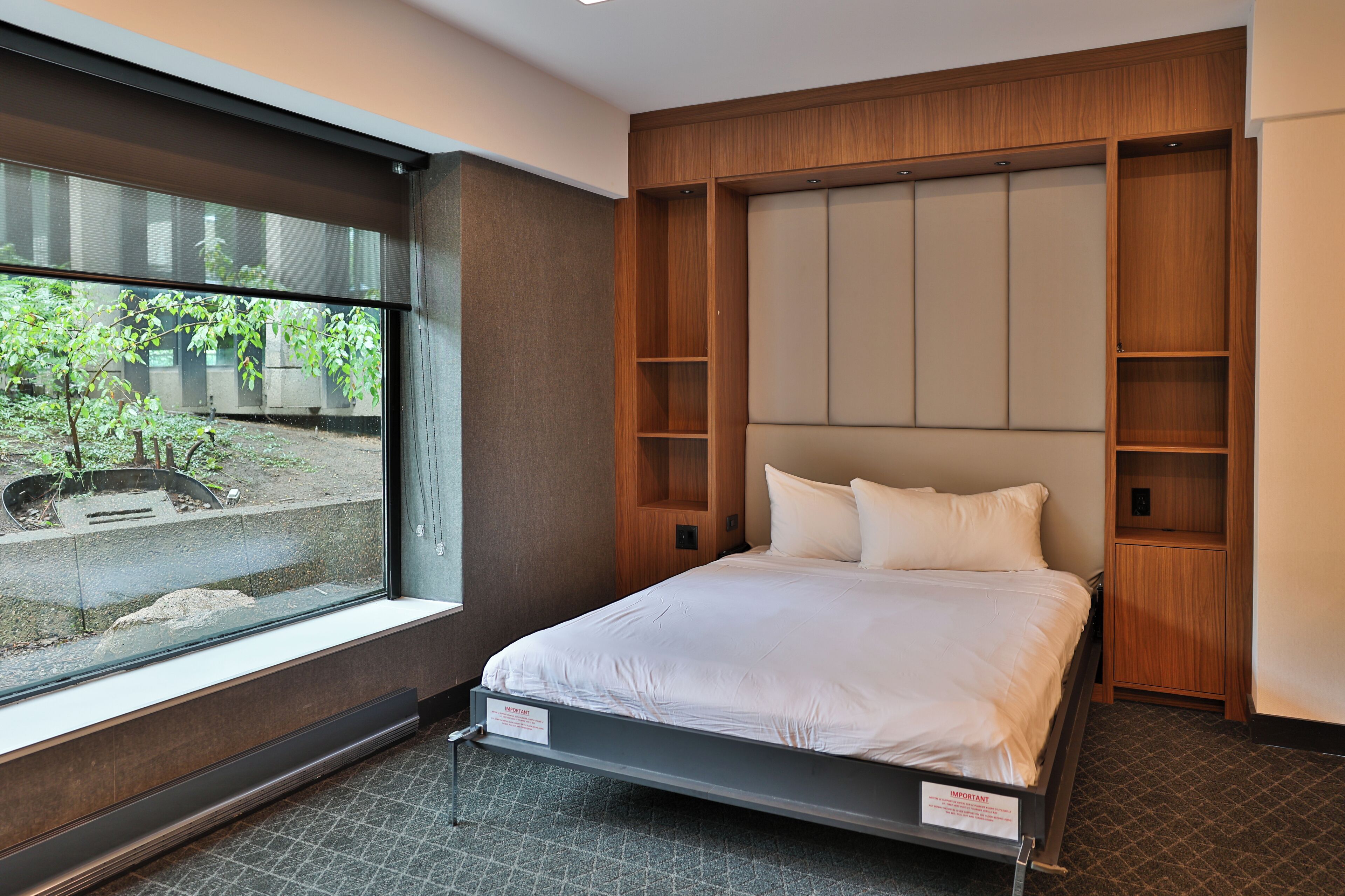 Une chambre d’hôtel moderne dotée d’une grande fenêtre, d’un lit avec du linge de maison blanc, de lambris en bois aux murs et d’étagères intégrées.