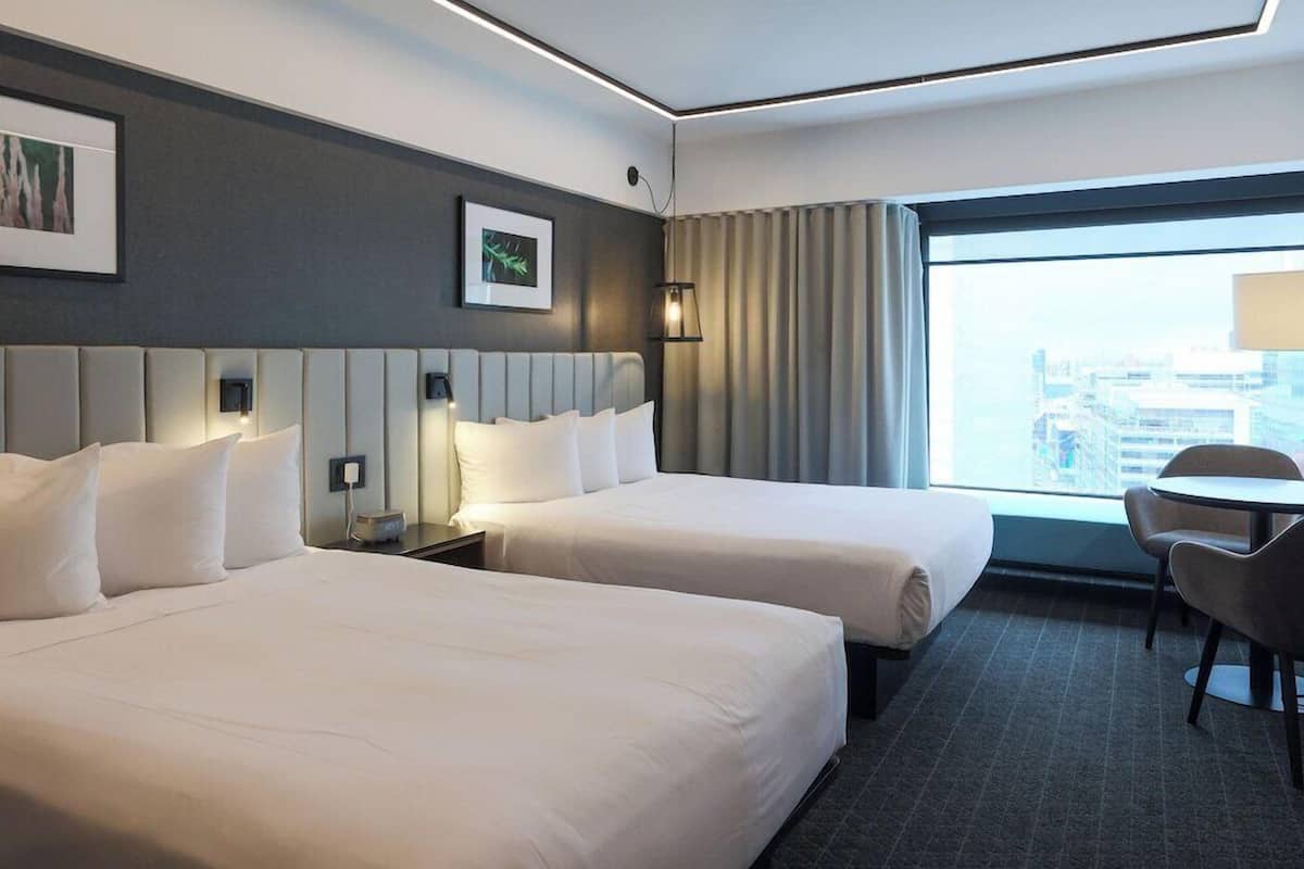 A hotel room with two beds, a desk, a chair, a city view, and a framed picture on the wall.