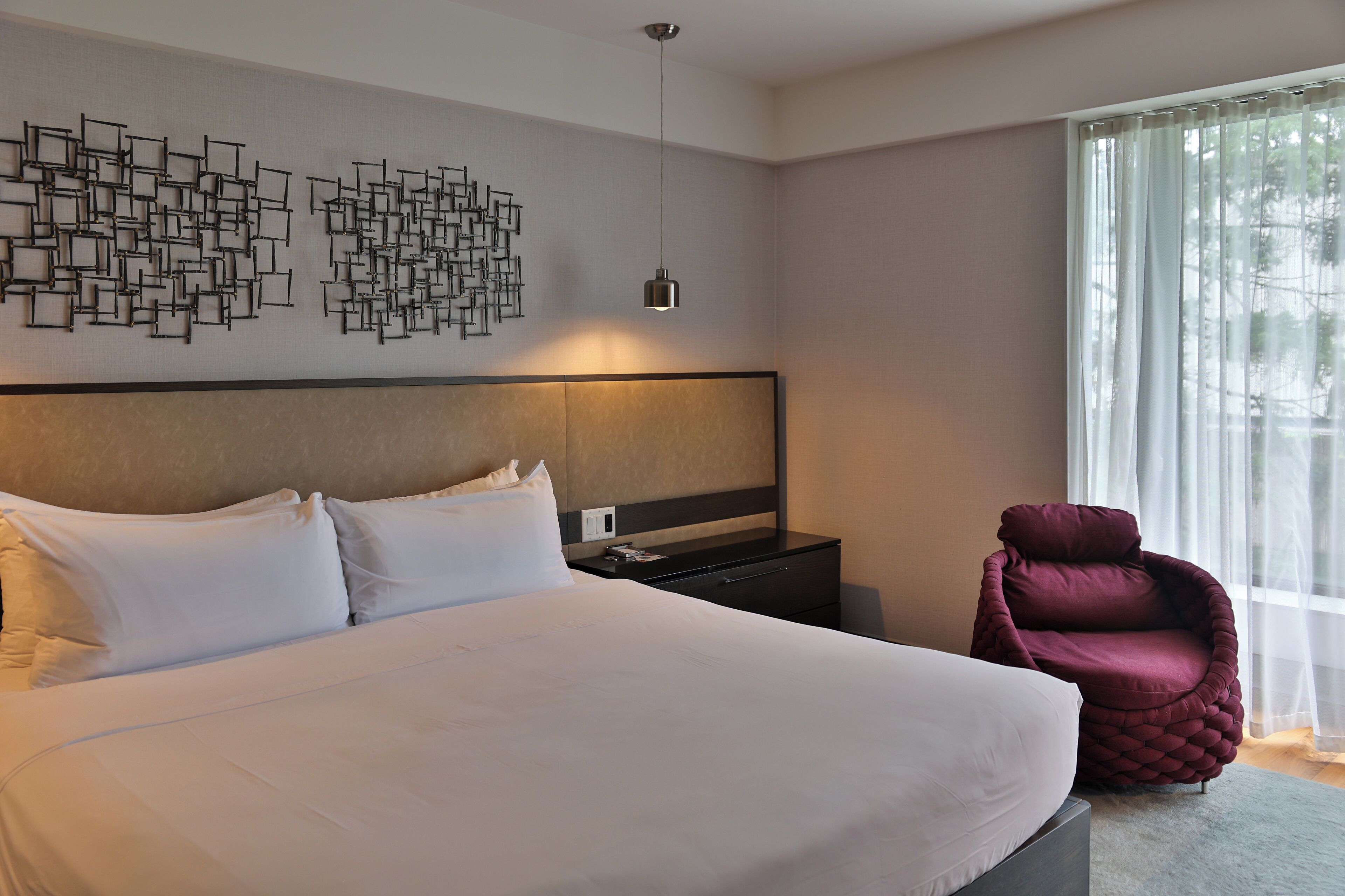 A modern hotel room with a large bed, a decorative wall art piece, a comfortable armchair, and a large window with sheer curtains.