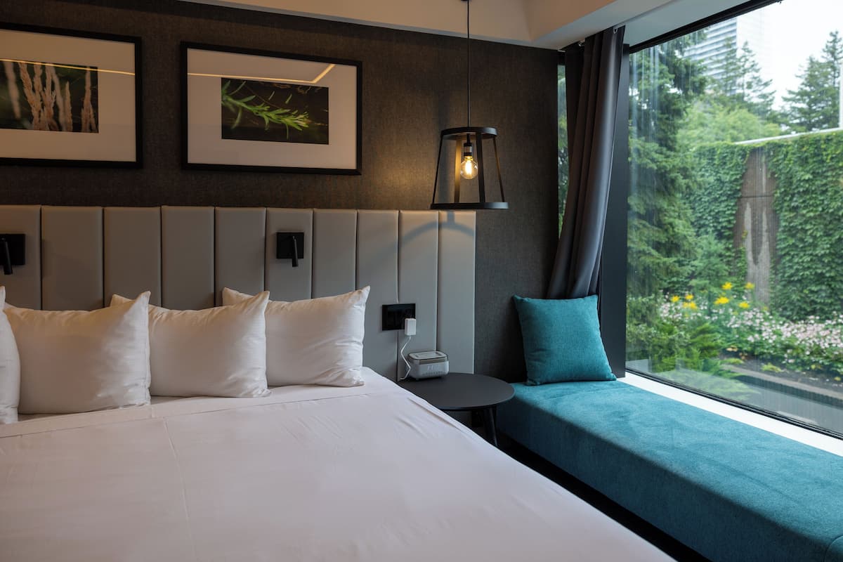 A modern hotel room with a large bed, a small bedside table, a lamp, and a window with a view of greenery.
