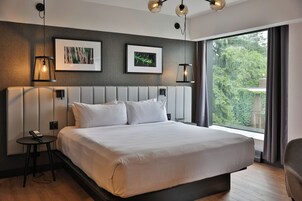 Una habitación de hotel moderna con una cama grande, dos cuadros colgados en la pared, una mesita de noche con un teléfono y un ventanal que ofrece vistas a la vegetación.