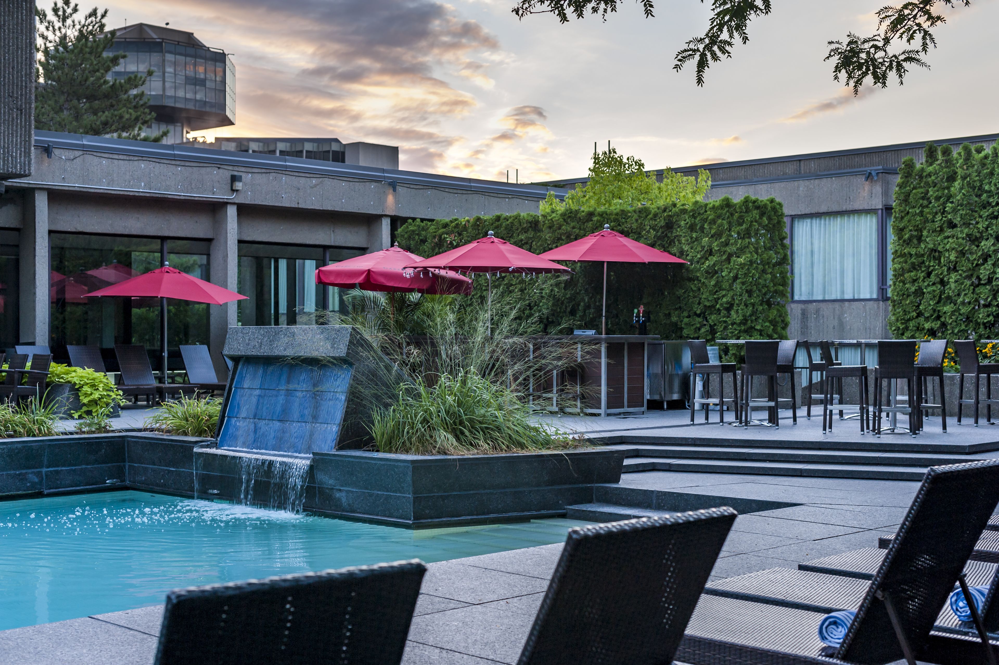 A pool area with lounge chairs and red umbrellas, surrounded by greenery and a modern building.