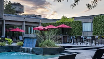 A pool area with lounge chairs and red umbrellas, surrounded by greenery and a modern building.