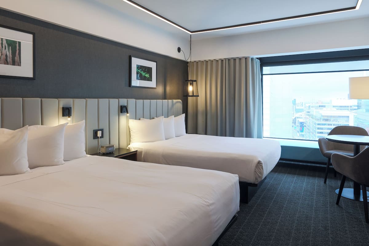 A hotel room with two beds, a desk, and a chair. There is a window with a view of buildings.