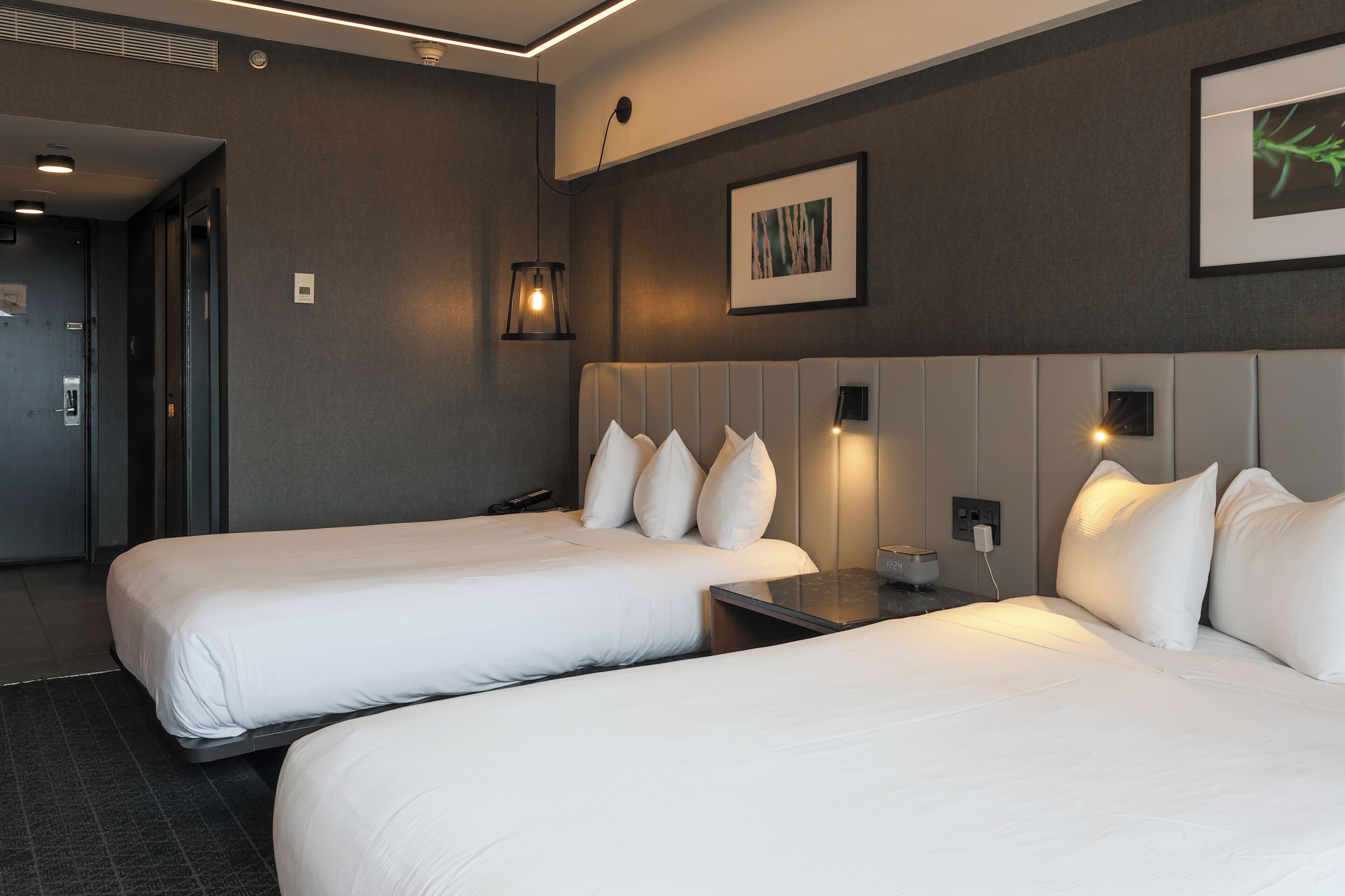 A hotel room with two beds, a bedside table, a wall-mounted lamp, and a framed picture.