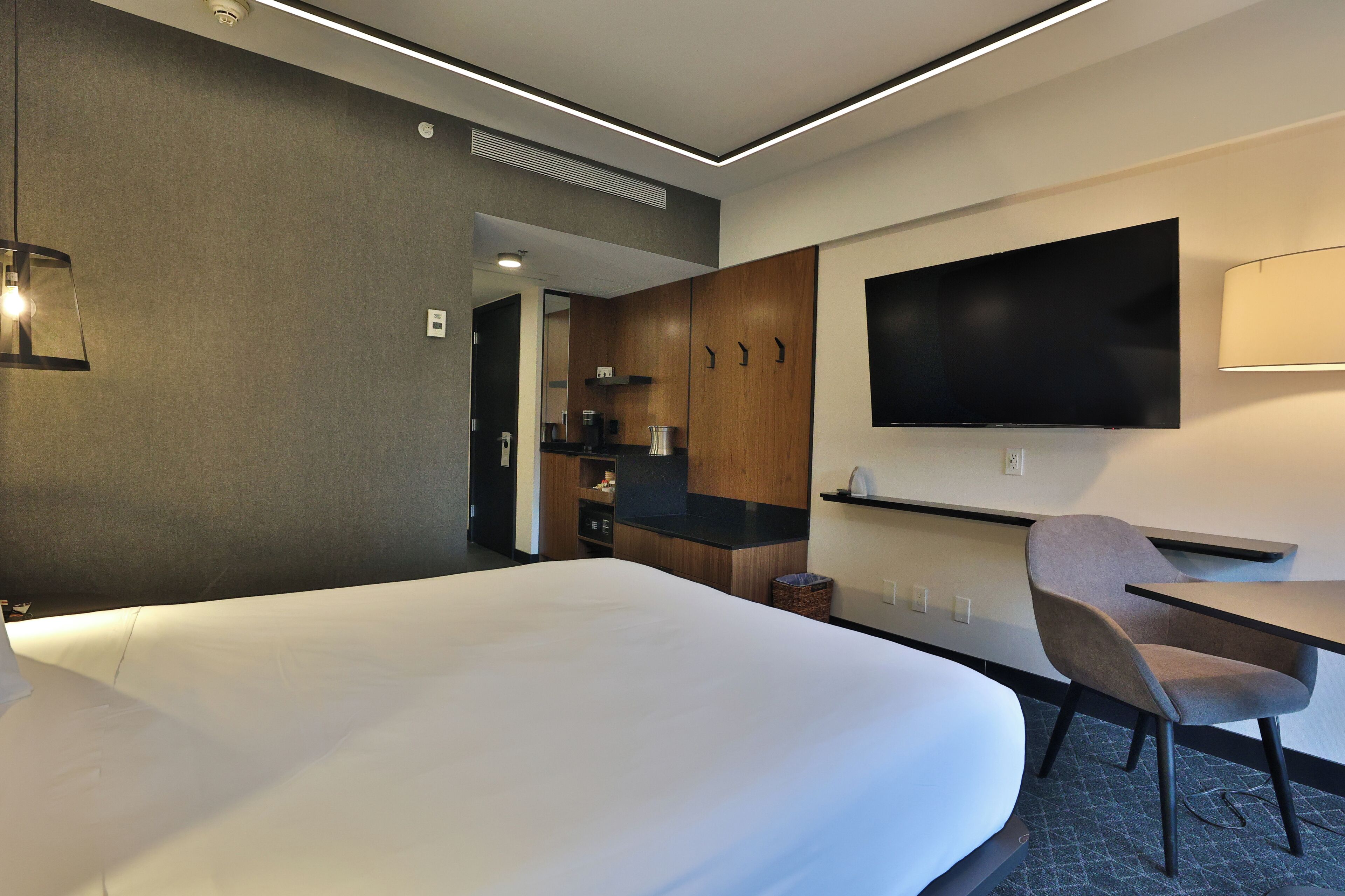 A hotel room with a large bed, a desk, a chair, a flat-screen TV mounted on the wall, and a kitchenette area.