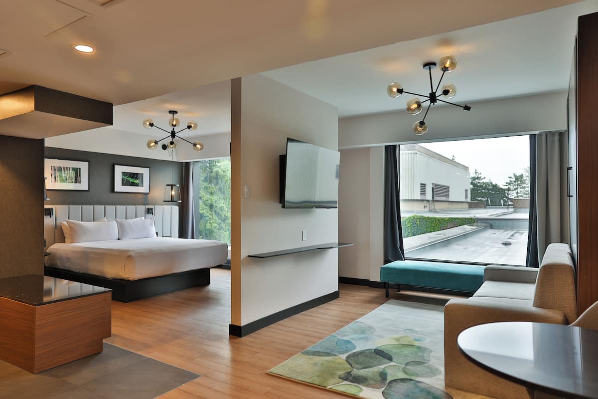 A modern hotel room with a bed, a desk, a sofa, a TV, and a large window with a view of the outside.
