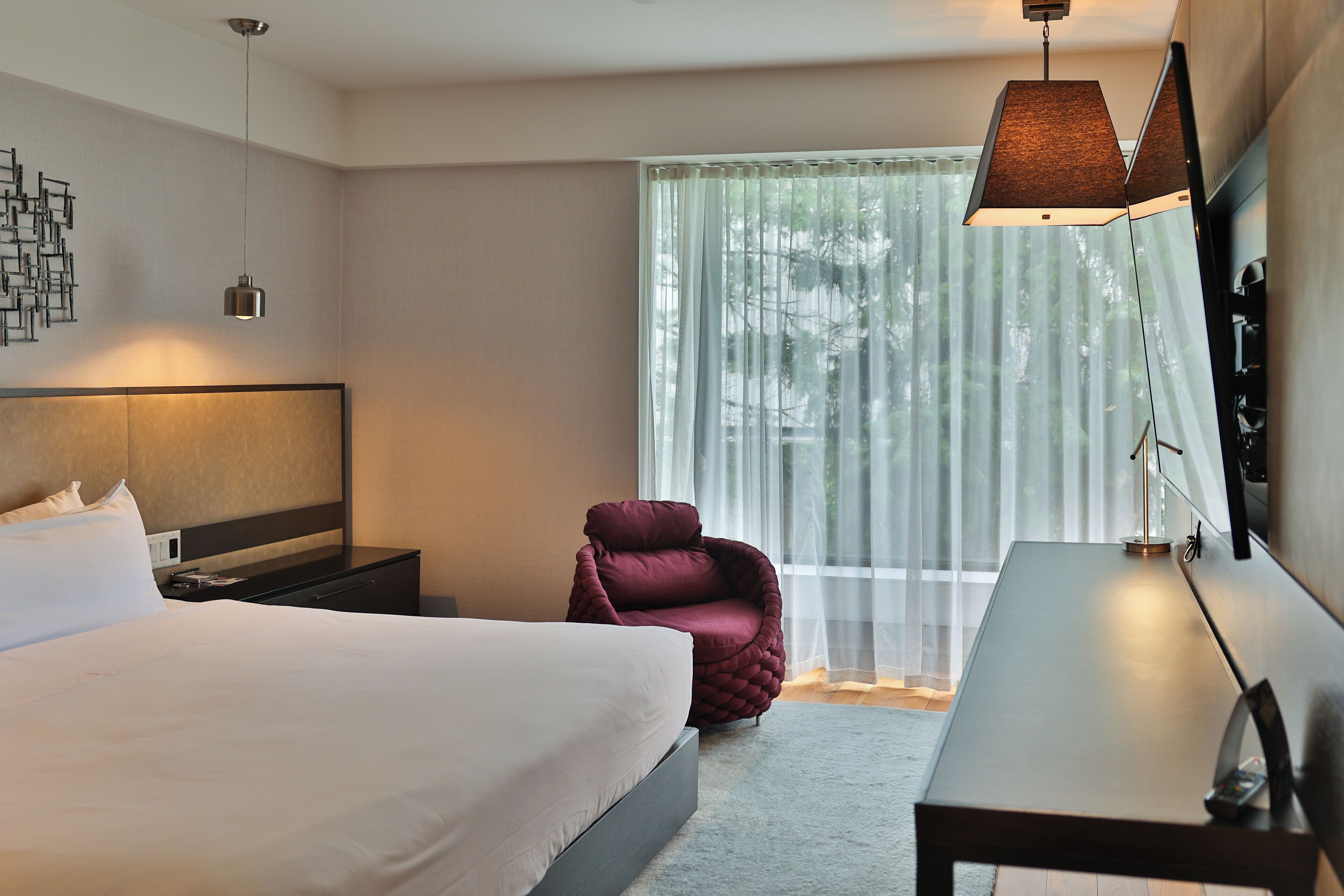 Une chambre d’hôtel moderne, dotée d’un grand lit, d’un bureau, d’une chaise et d’une grande fenêtre avec des rideaux légers.