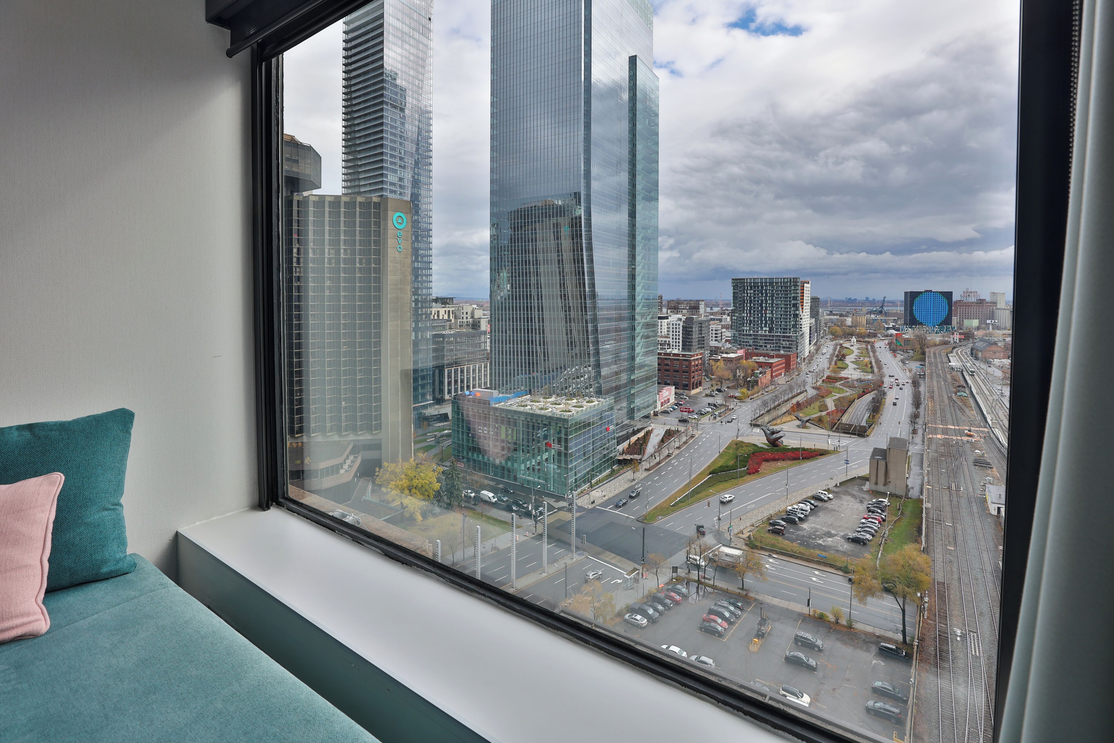 Una habitación con un amplio ventanal que ofrece vistas a un paisaje urbano con edificios altos y un estacionamiento.