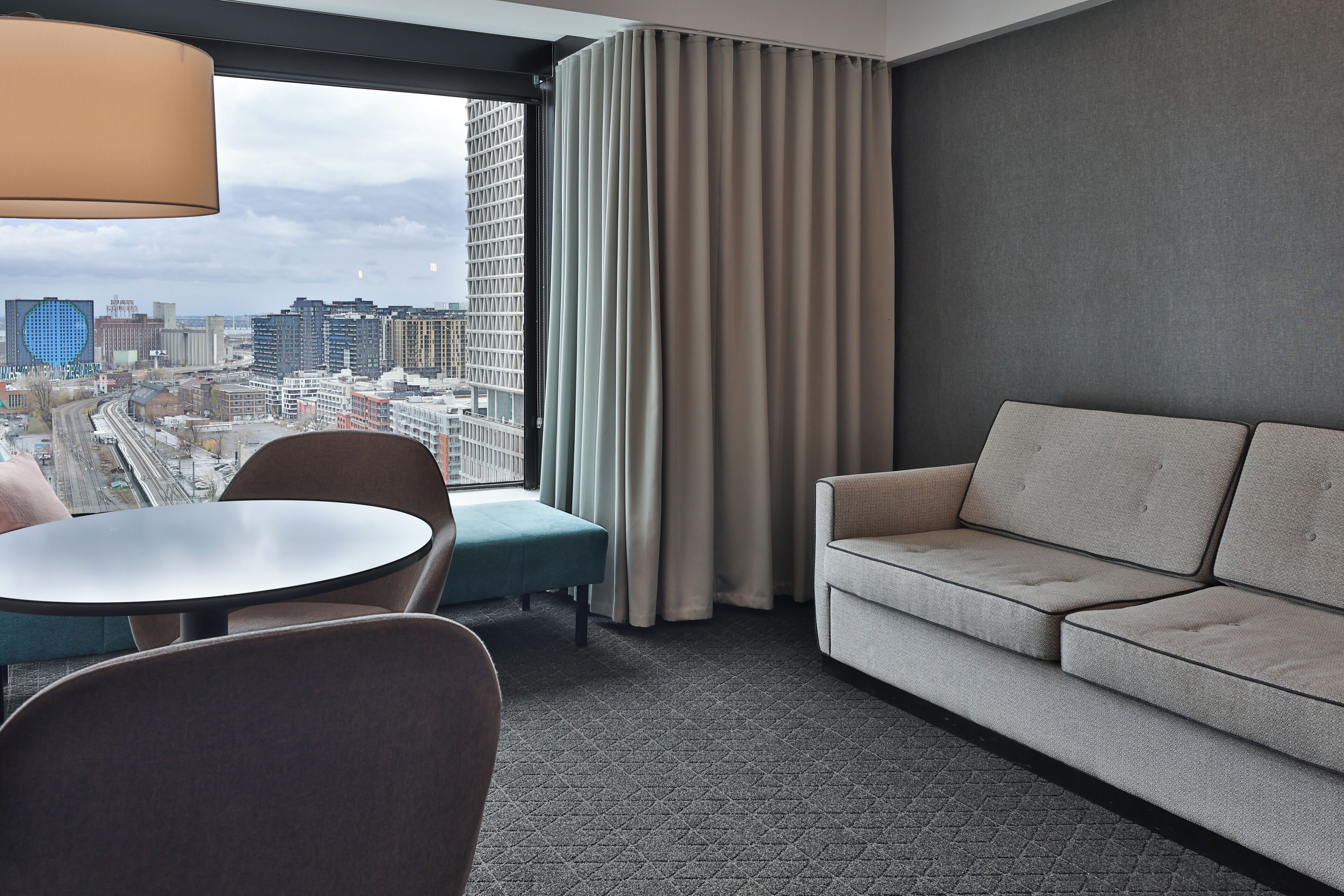 A hotel room with a large window, a sofa, a round table, and a chair.