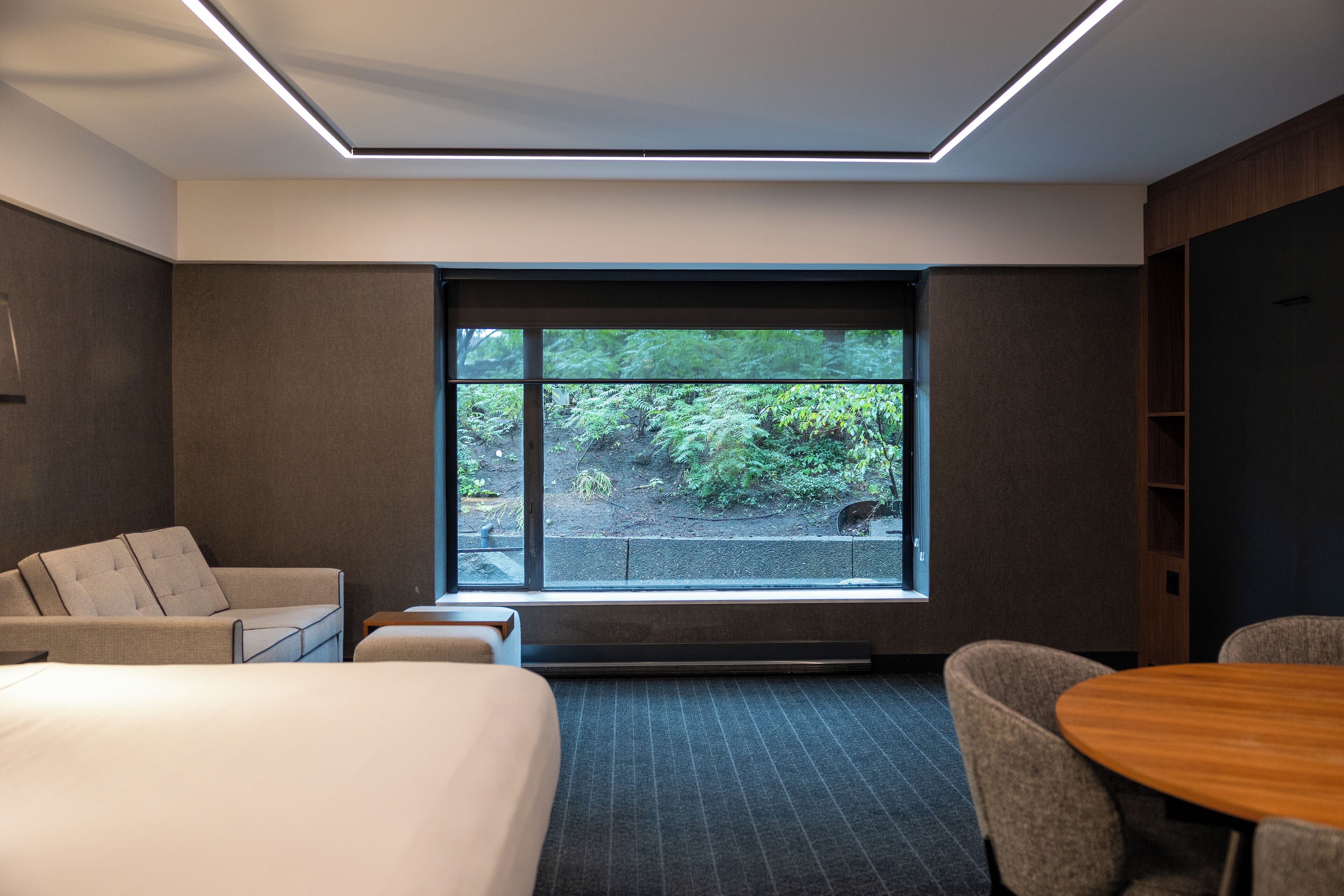 A modern hotel room with a large window, a bed, a sofa, a table, and a chair.