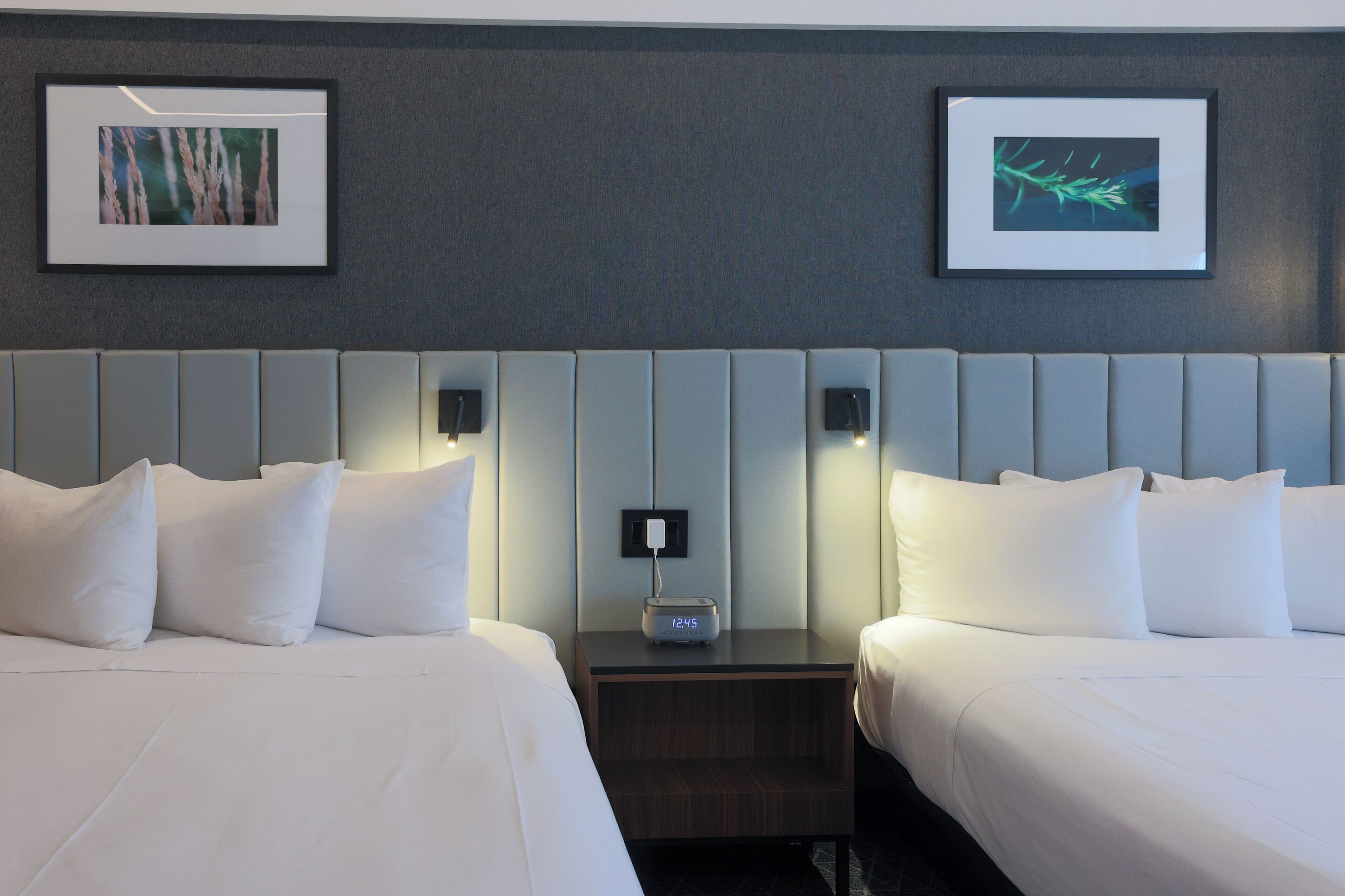 A hotel room with two beds, a nightstand with a clock, and framed artwork on the wall.