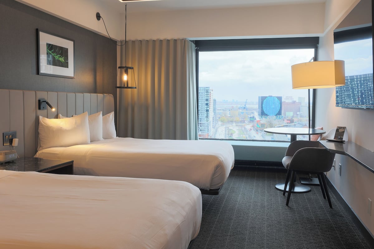 A hotel room with two beds, a desk, a chair, and a large window offering a city view.
