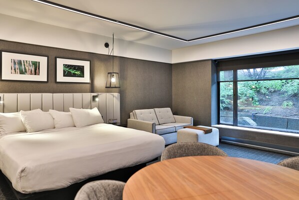 Una habitación de hotel moderna con una cama grande, un sofá, una mesa de comedor y un ventanal con vistas a zonas verdes.