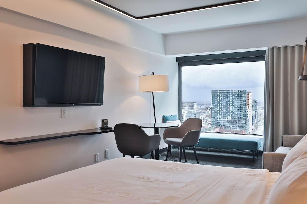 A hotel room with a bed, two chairs, a small table, a lamp, a TV, and a large window with a city view.