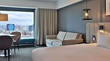 A hotel room with a bed, a sofa, a chair, a desk, a lamp, and a view of the cityscape.