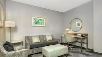 Una habitación de hotel con un sofá gris, un sillón a juego, un escritorio pequeño con una silla, una lámpara y una obra de arte enmarcada en la pared.
