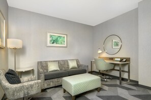 Une chambre d’hôtel avec un canapé gris, un fauteuil assorti, un petit bureau avec une chaise, une lampe et une œuvre d’art encadrée accrochée au mur.