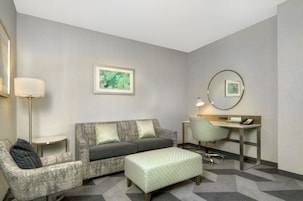 A hotel room with a grey sofa, a matching armchair, a small desk with a chair, a lamp, and a framed artwork on the wall.